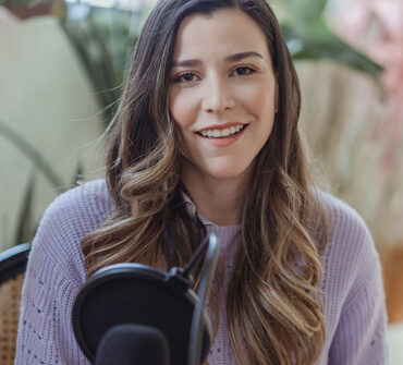 Smiling female audio host in a cozy setting, wearing a lavender sweater, with a microphone in front, representing Blueprints and Beginnings' focus on audio content.