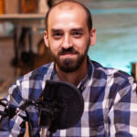 Mike Hardson Podcast host with microphone, wearing a plaid shirt, smiling in a creative studio setting, reflecting the theme of lifestyle and fashion discussions in the podcast episode "Everything looks so tiny from up here."