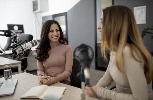 Two women engaged in a podcast discussion, one smiling and holding a pen, seated at a table with a laptop and microphone, in a modern studio setting.