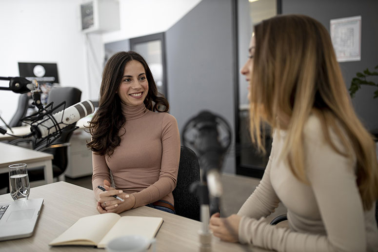 Two women engaging in conversation at a podcast recording studio, one smiling while holding a pen and notebook, with microphones and a laptop on the table.