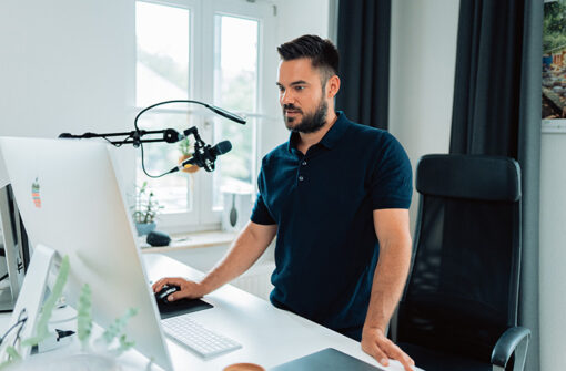 Man standing at a desk with a computer and microphone, engaged in podcasting or audio recording, relevant to the "Everything looks so tiny from up here" episode on lifestyle and fashion.