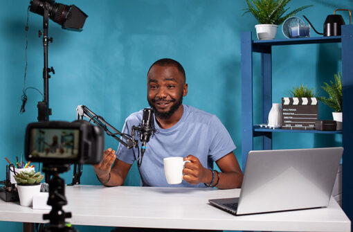 Man sitting at a desk with a microphone and laptop, holding a cup, in a podcast recording setup against a blue background, representing audio content creation for the 'Multiple Media' category.