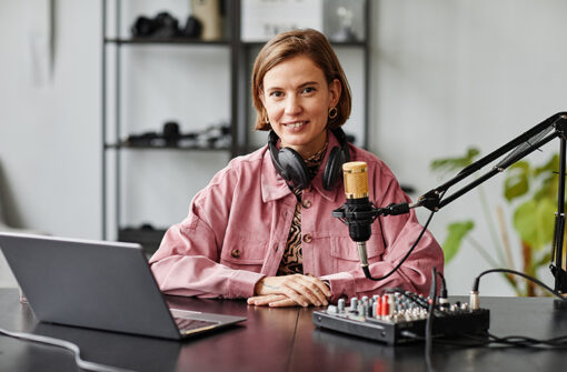 Woman in a pink jacket with headphones sitting at a desk with a microphone and audio mixer, representing podcast production for 'This is Where You'll Find the Best Pool Accessories'.