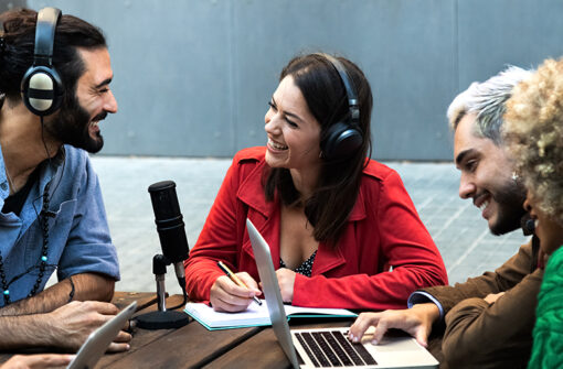Group of podcast hosts discussing topics related to fashion and lifestyle, with microphones, laptops, and notebooks at a table.