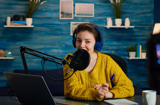 Woman in a yellow sweater wearing headphones, speaking into a microphone in a podcasting setup with a laptop and notepad, reflecting the theme of engaging audio content.