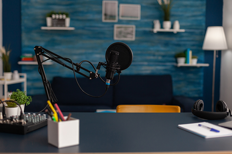 Podcasting setup with a microphone on an adjustable arm, audio mixer, and stationery on a desk, featuring a blue wall, couch, and decorative shelves, reflecting a creative workspace for discussing website design using Figma.