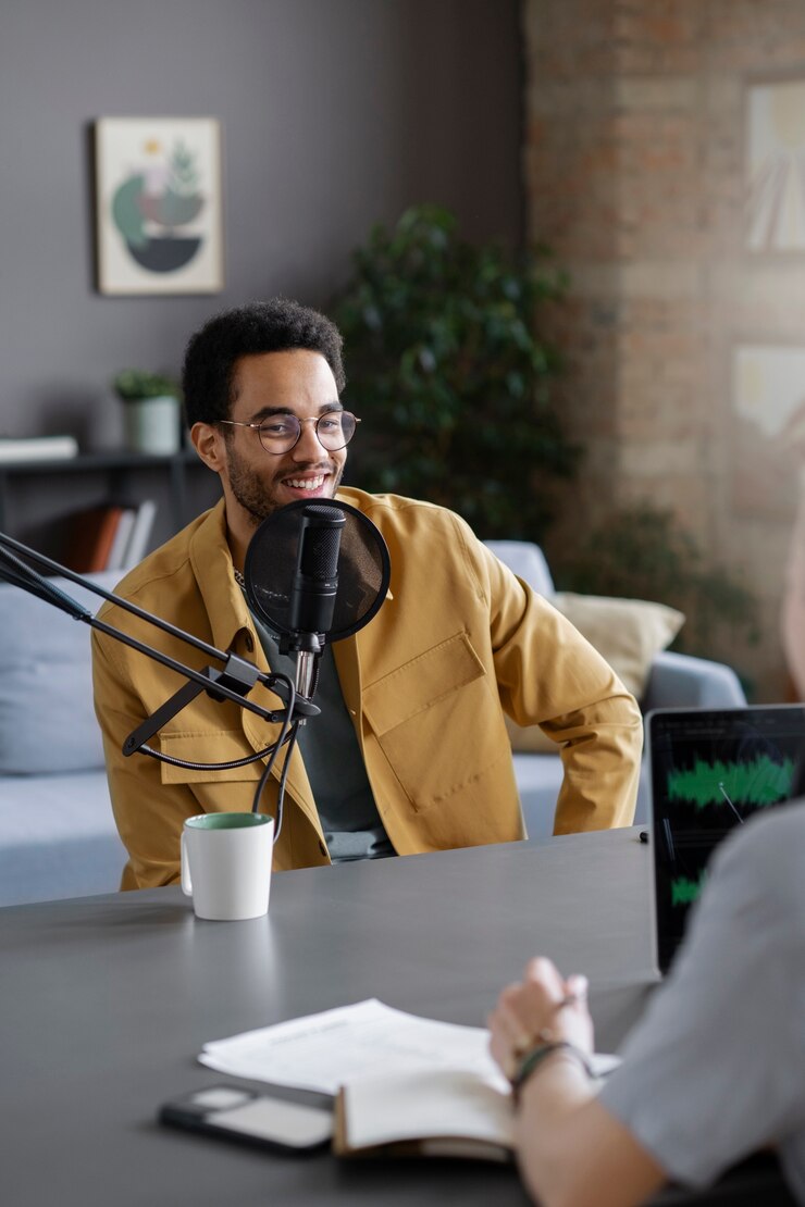 Smiling man in casual attire speaking into a microphone during a podcast recording, with a laptop and notebook on the table, reflecting themes of mentorship and career development from the Blueprints & Beginnings podcast.